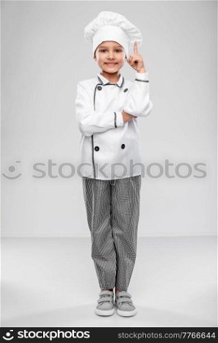 cooking, culinary and profession concept - happy smiling little girl in chef&rsquo;s toque and jacket pointing finger up over grey background. little girl in chef&rsquo;s toque pointing finger up