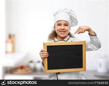 cooking, culinary and profession concept - happy smiling little girl in chef&rsquo;s toque and jacket holding chalkboard over kitchen background. happy little girl in chef&rsquo;s toque with chalkboard