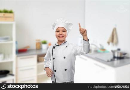 cooking, culinary and profession concept - happy smiling little girl in chef&rsquo;s toque and jacket pointing finger up over home kitchen background. girl in chef&rsquo;s toque pointing finger up in kitchen