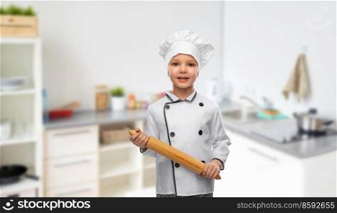 cooking, culinary and profession concept - happy smiling little girl in chef’s toque and jacket with rolling pin over home kitchen background. girl in chef’s toque with rolling pin in kitchen