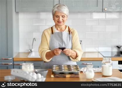 cooking, food and culinary concept - happy smiling senior woman with smartphone taking picture of cupcakes in baking mold on kitchen at home. woman with smartphone and cupcakes on kitchen