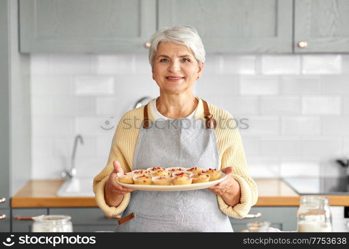 cooking food, baking and culinary concept - happy smiling senior woman holding big plate with cupcakes on kitchen at home. woman holding plate with cupcakes on kitchen