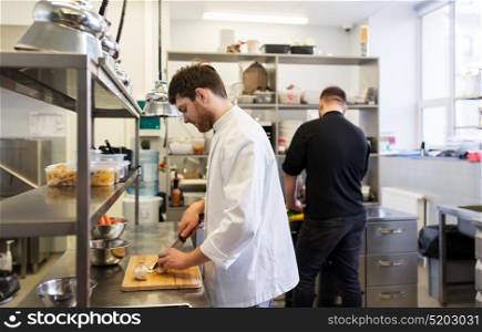 cooking food, profession and people concept - happy male chef and cook with knife chopping champignons on cutting board at restaurant kitchen. chef and cook cooking food at restaurant kitchen