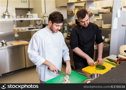 cooking food, profession and people concept - happy male chef and cook with knife chopping champignons and greens on cutting boards at restaurant kitchen. chef and cook cooking food at restaurant kitchen