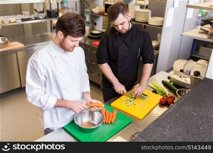cooking food, profession and people concept - happy male chef cook with knife peeling carrot and chopping cucumber on cutting board at restaurant kitchen. happy male chef cooking food at restaurant kitchen