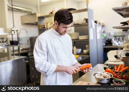 cooking food, profession and people concept - happy male chef cook with knife peeling carrot on cutting board at restaurant kitchen. happy male chef cooking food at restaurant kitchen