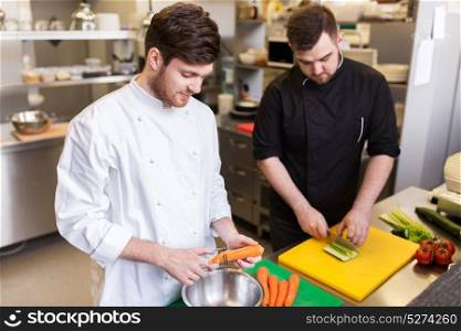 cooking food, profession and people concept - happy male chef cook with knife peeling carrot and chopping cucumber on cutting board at restaurant kitchen. chef and cook cooking food at restaurant kitchen