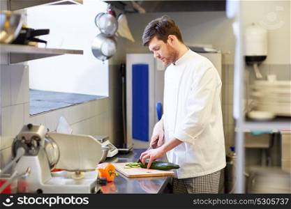 cooking food, profession and people concept - happy male chef cook with knife chopping cucumber on cutting board at restaurant kitchen. happy male chef cooking food at restaurant kitchen