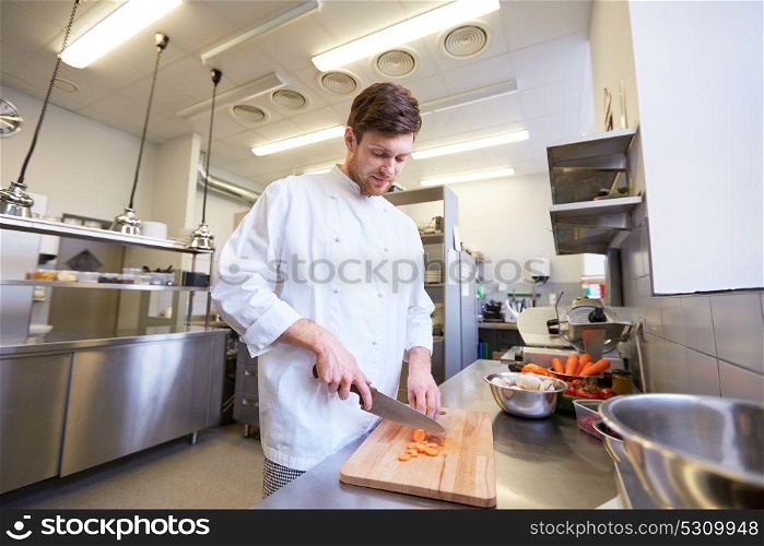 cooking food, profession and people concept - happy male chef cook with knife chopping carrot on cutting board at restaurant kitchen. happy male chef cooking food at restaurant kitchen