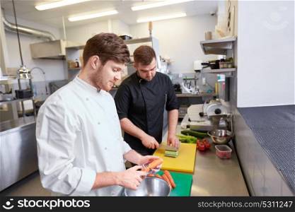 cooking food, profession and people concept - happy male chef cook with knife peeling carrot and chopping cucumber on cutting board at restaurant kitchen. chef and cook cooking food at restaurant kitchen