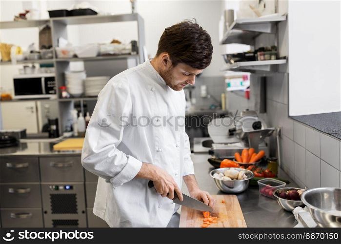 cooking food, profession and people concept - happy male chef cook with knife chopping carrot on cutting board at restaurant kitchen. happy male chef cooking food at restaurant kitchen. happy male chef cooking food at restaurant kitchen