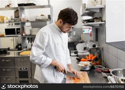 cooking food, profession and people concept - happy male chef cook with knife chopping carrot on cutting board at restaurant kitchen. happy male chef cooking food at restaurant kitchen. happy male chef cooking food at restaurant kitchen