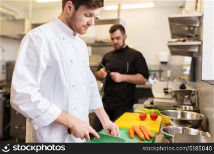 cooking food, profession and people concept - happy male chef with knife chopping carrot on cutting board and cook sharpening knife at restaurant kitchen. chef and cook cooking food at restaurant kitchen