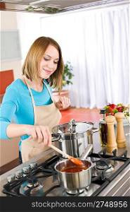 Cooking - Happy woman by stove in kitchen with pots and pans stirring tomato sauce
