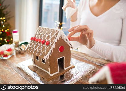 cooking, holidays and people concept - woman with pastry bag making gingerbread house at home over christmas tree lights background. close up of woman making gingerbread house