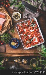Cooking in a vintage kitchen. Tomatoes on a baking sheet, raw fish , herbs and seasonings, olives oil , cleaver on a rustic wooden table background. Top view. Mediterranean food concept