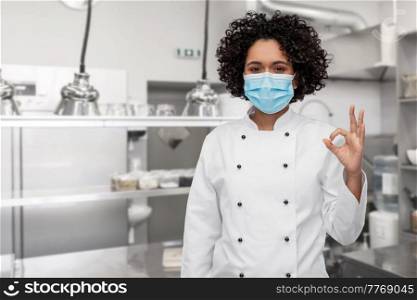 cooking, pandemic and health concept - happy smiling female chef in protective medical mask and white jacket showing ok gesture over restaurant kitchen background. female chef in medical mask showing ok on kitchen