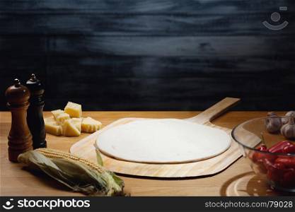 Cooking pizza background, dough base on board. Low key shot, light on dough, some ingredients around on table. Copy space.
