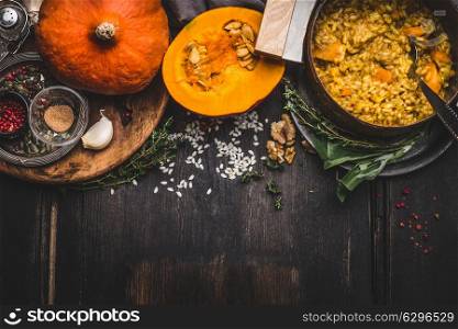 Cooking pot of vegetarian pumpkin risotto and spoon on dark rustic kitchen table background with cooking ingredients, top view, border. Healthy clean seasonal food and eating concept