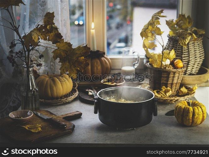 Cooking pot with steam and millet on kitchen table with pumpkins, autumn leaves, candles and kitchen utensils at window background. Cooking healthy grains at home. Front view.