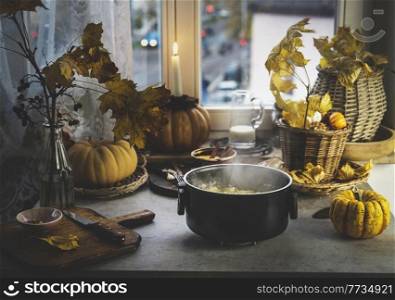 Cooking pot with steam and millet on kitchen table with pumpkins, autumn leaves, candles and kitchen utensils at window background. Cooking healthy grains at home. Front view.