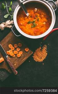 Cooking pot with tasty lentil soup and ladle on dark rustic kitchen table background with ingredients, top view with copy space. Healthy vegan food concept