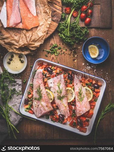 Cooking preparation of Bass fish fillets in Mediterranean sauce with tomatoes, olives and capers in baking pan on rustic wooden background with ingredients. Top view