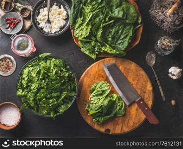 Cooking preparation of delicious vegetarian spinach dish. Cutting board with knife on rustic kitchen background. Healthy ingredients are laid out around. Top view.