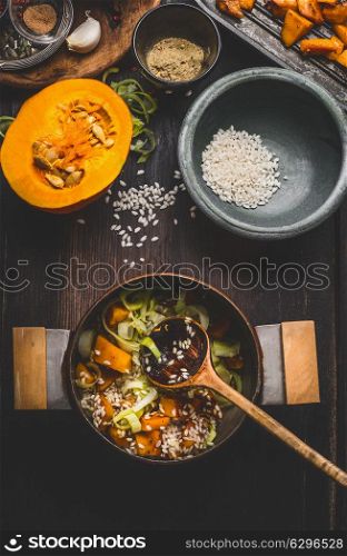 Cooking preparation with pot of vegetarian pumpkin risotto and spoon on dark rustic kitchen table background with cooking ingredients, top view. Healthy clean seasonal food and eating concept