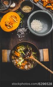 Cooking preparation with pot of vegetarian pumpkin risotto and spoon on dark rustic kitchen table background with cooking ingredients, top view. Healthy clean seasonal food and eating concept