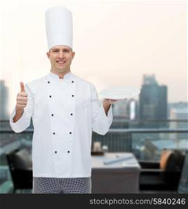 cooking, profession, advertisement and people concept - happy male chef cook holding something on empty plate and showing thumbs up over city restaurant lounge background