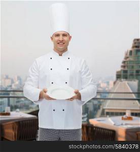 cooking, profession, advertisement and people concept - happy male chef cook showing something on empty plate over city restaurant lounge background