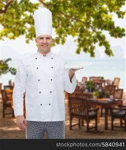 cooking, profession, advertisement and people concept - happy male chef cook showing something on empty plate over restaurant lounge on beach