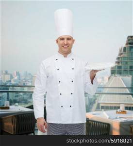 cooking, profession, advertisement and people concept - happy male chef cook showing something on empty plate over city restaurant lounge background