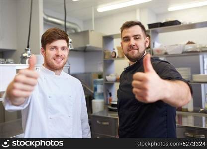 cooking, profession and people concept - happy male chef and cook at restaurant kitchen showing thumbs up. chefs at restaurant kitchen showing thumbs up