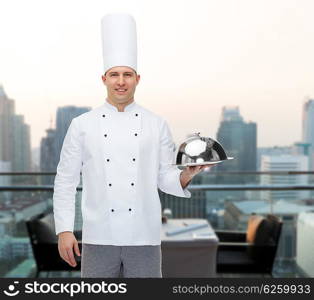 cooking, profession and people concept - happy male chef cook holding cloche over city restaurant lounge background