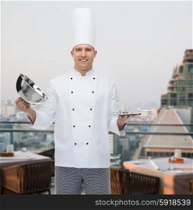 cooking, profession and people concept - happy male chef cook opening cloche cover over city restaurant lounge background