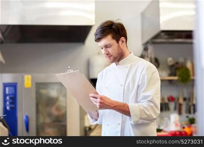 cooking, profession and people concept - happy male chef cook writing to clipboard and doing inventory at restaurant kitchen. chef with clipboard doing inventory at restaurant