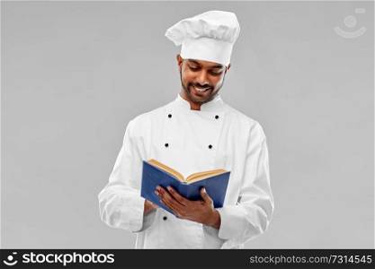 cooking, profession and people concept - happy male indian chef in toque reading cookbook over grey background. happy male indian chef reading cookbook