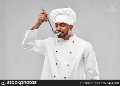 cooking, profession and people concept - happy male indian chef in toque tasting food from ladle over grey background. happy male indian chef tasting food from ladle