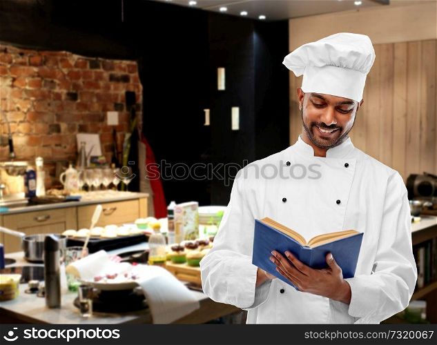 cooking, profession and people concept - happy male indian chef in toque reading cookbook over restaurant kitchen background. happy male indian chef reading cookbook at kitchen