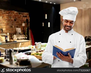 cooking, profession and people concept - happy male indian chef in toque reading cookbook over restaurant kitchen background. happy male indian chef reading cookbook at kitchen
