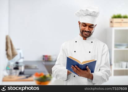 cooking, profession and people concept - happy male indian chef in toque reading cookbook over kitchen background. happy male indian chef reading cookbook at kitchen