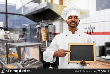 cooking, profession and people concept - happy male indian chef in toque with blank chalkboard for menu over kebab shop kitchen background. happy indian chef with chalkboard at kebab shop