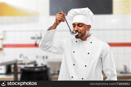 cooking, profession and people concept - happy male indian chef in toque tasting food from ladle over restaurant kitchen background. happy indian chef tasting food by ladle at kitchen