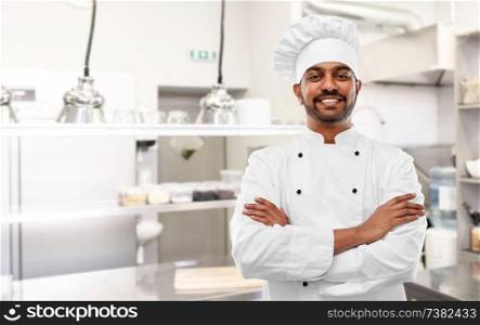 cooking, profession and people concept - happy male indian chef in toque with crossed arms over restaurant kitchen background. happy indian chef in toque at restaurant kitchen