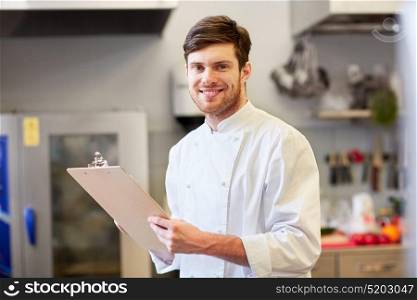 cooking, profession and people concept - happy smiling male chef cook writing to clipboard and doing inventory at restaurant kitchen. chef with clipboard doing inventory at restaurant