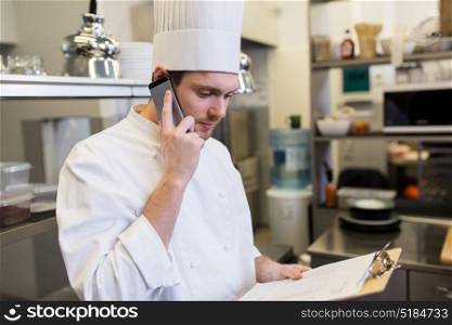 cooking, profession and people concept - male chef cook with clipboard calling on smartphone at restaurant kitchen. chef calling on smartphone at restaurant kitchen