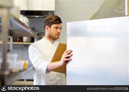 cooking, profession and people concept - male chef cook with clipboard doing inventory and looking to fridge at restaurant kitchen. chef with clipboard doing inventory at kitchen
