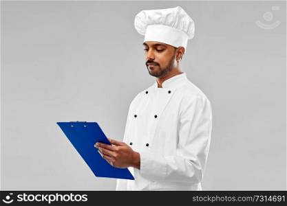 cooking, profession and people concept - male indian chef in toque reading menu on clipboard over grey background. male indian chef reading menu on clipboard